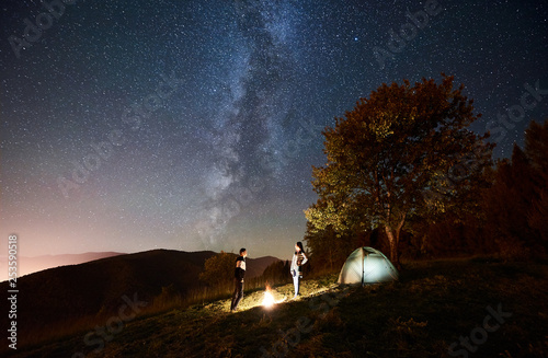 Young happy couple tourists standing at bonfire near illuminated tent under amazing night sky full of stars and Milky way. On the background beautiful starry sky  mountains and big tree