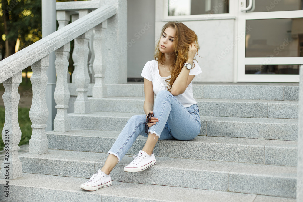 Modern attractive stylish young woman in a fashionable t-shirt in blue jeans  in white sneakers resting sitting on the steps of a vintage building. Cute  European girl enjoying summer day outdoors. Stock