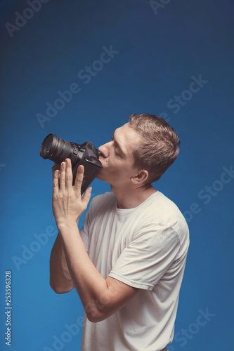 Smiling Man with camera isolated on blue background. Young man holding digital camera and kiss it. Lifestyle, travel and technology concept. Happy boy in white t-shirt love his dslr camera