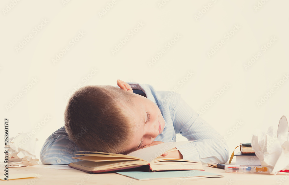 Cute little school boy with huge smile sitting at his desk on white background. Happy intelligent children in shirt with blue eyes, lies on table with book and sleep Cute little school boy with huge smile sitting at his desk on white background. Happy intelligent children in shirt with blue eyes, lies on table with book and sleep