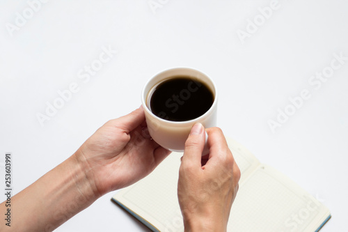 Female hands hold a cup of black coffee and an open diary with clean pages, a pen on a light background. Minimalist style and creative concepts. Flat lay, top view