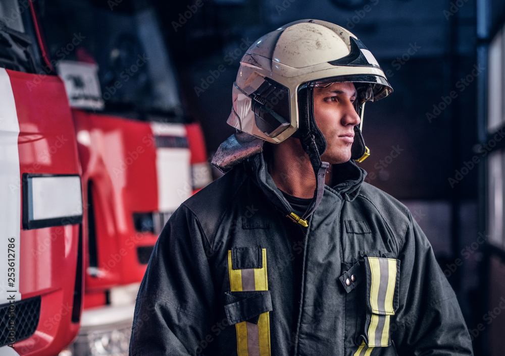 Brave young fireman wearing protective uniform standing next to a fire engine in a garage of a fire department and looking outside Brave young fireman wearing protective uniform standing next to a fire engine in a garage of a fire department and looking outside