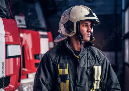 Brave young fireman wearing protective uniform standing next to a fire engine in a garage of a fire department and looking outside
