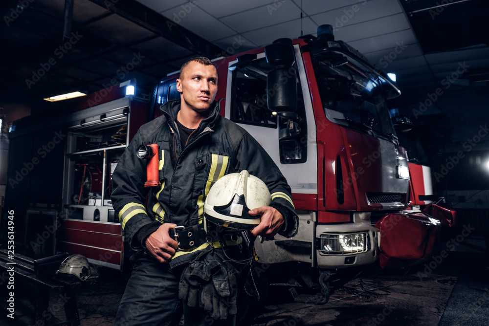 Fireman wearing uniform holding a helmet and looking at a camera while standing near a fire truck in a garage of a fire department at night. Fireman wearing uniform holding a helmet and looking at a camera while standing near a fire truck in a garage of a fire department at night.