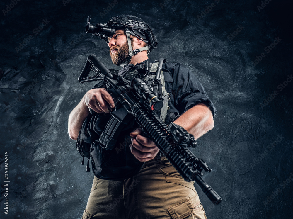 Bearded special forces soldier or private military contractor holding an assault rifle and observes the surroundings in night vision goggles. Studio photo against a dark textured wall