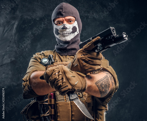 A terrorist in a military uniform and a skull balaclava holding a pistol and a knife and looks at the camera with a menacing look. Studio photo against a dark textured wall