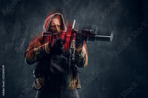 Special forces soldier in military uniform wearing mask and hood holding an assault rifle with a laser sight and aims at the target. Studio photo against a dark textured wall