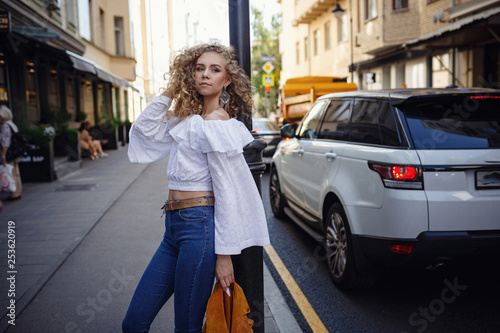 A beautiful young woman with blond curly hair walks through the sunny city