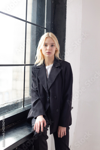 young beautiful girl, posing for a photo on a studio near a large window dressed in a black trouser suit with a jacket, office modern style