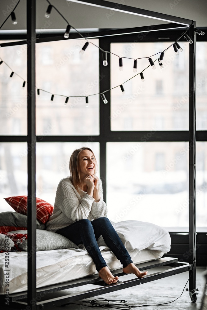 A beautiful girl in blue jeans and a white sweater posing in a metal cube and at the window.