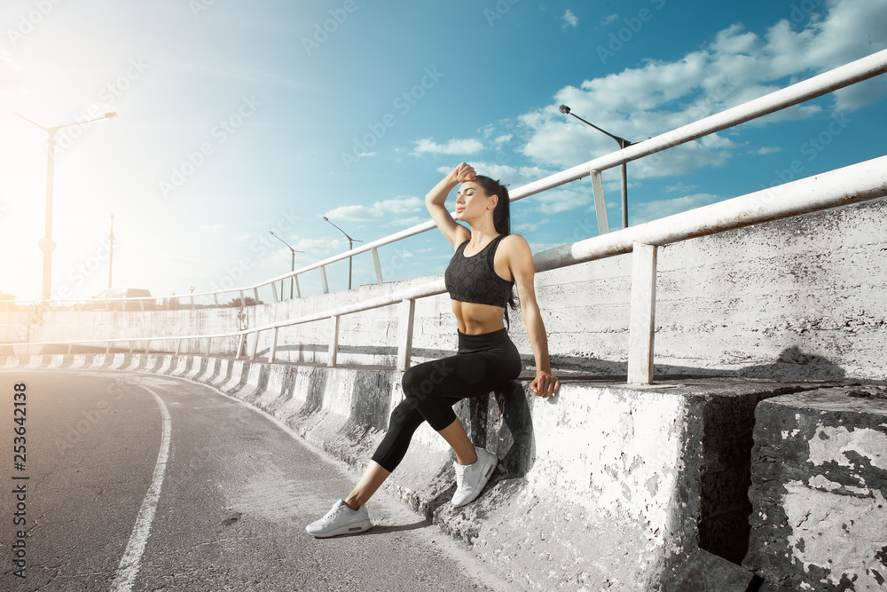 Fototapeta premium Portrait of beautiful brunette. Sexy girl rest after running. Female fitness model working out outdoor in city. Young woman jogger athlete training on road. Concept of healthy lifestyle.