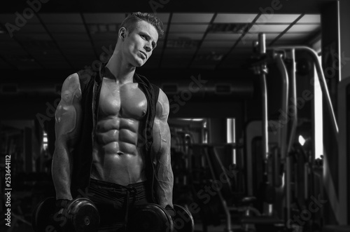 Muscular model sports young man exercising in gym with dumbbell. Black and white portrait of strong muscle. Fitness trainer. Sport workout bodybuilding motivation concept. Sexy torso.