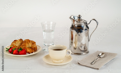 Cream Colored Plate and Coffee Cup with Two Croissants and Three Fresh Strawberries on a White Linen Tablecloth and a Silver Hotel Room Service Carafe with a Napkin and Spoon.