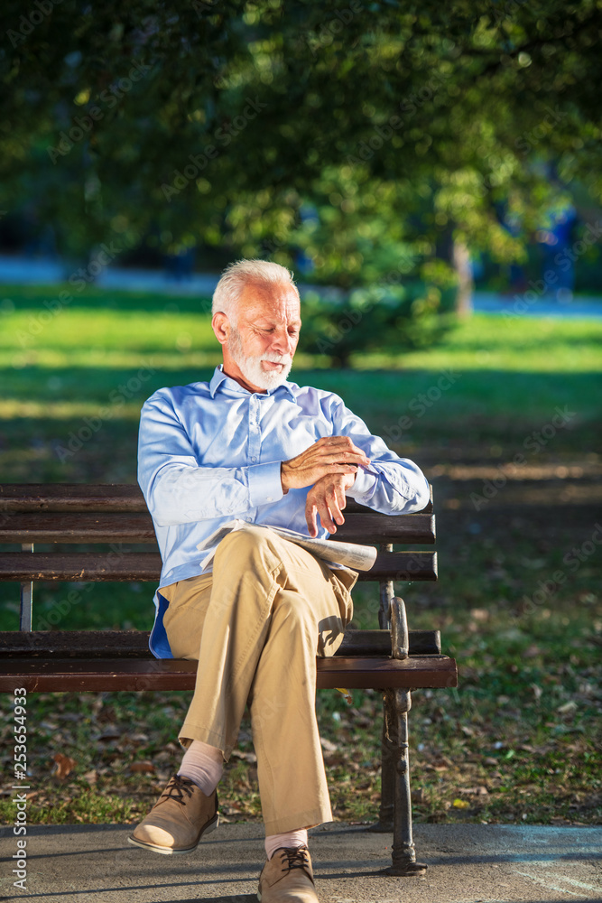 Fototapeta premium Senior man relaxing in park on a sunny day seated on a wooden bench and waiting for someone