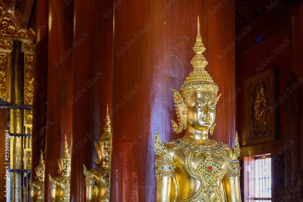 Row of golden Buddha statue statues stand in front of red columns inside Thai temple. 