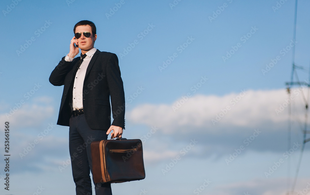 Handsome young businessman in a suit and tie with a suitcase is standing on a blue sky background and talking on a mobile phone. Sunglasses. Handsome young businessman in a suit and tie with a suitcase is standing on a blue sky background and talking on a mobile phone. Sunglasses.