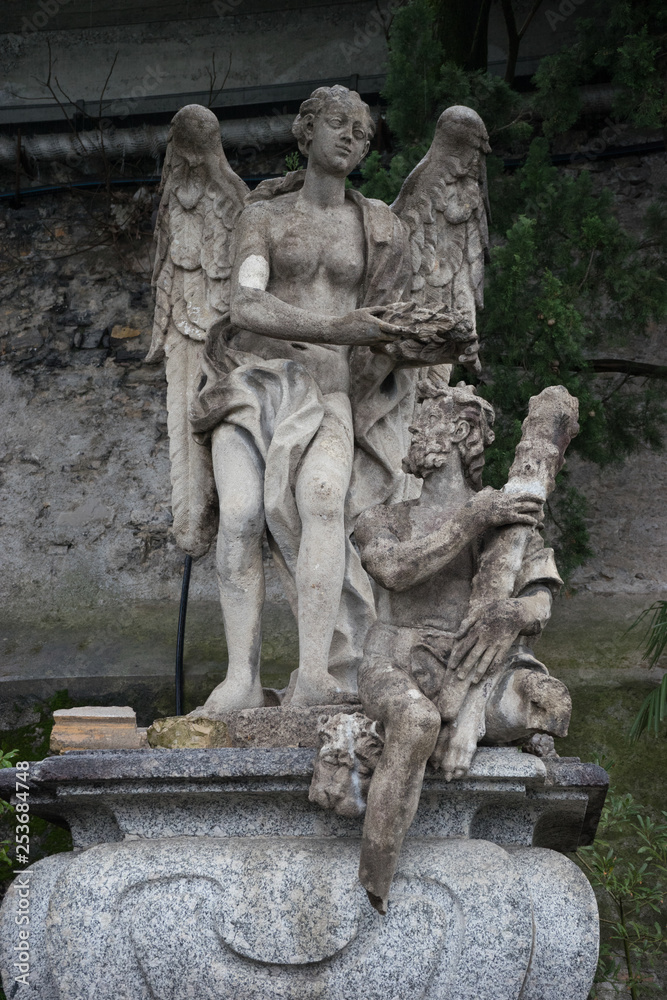Italy, Varenna, Lake ComoVarenna, Italy - March 31, 2018: Stone Sculpture artifact at gardens of Villa Monastero, a person sitting on a rock Italy, Varenna, Lake ComoVarenna, Italy - March 31, 2018: Stone Sculpture artifact at gardens of Villa Monastero, a person sitting on a rock