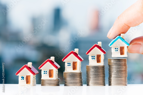 Concept for property ladder, mortgage and real estate investment. Man's hand putting blue house model on top of coins stack with city backgrounds. photo