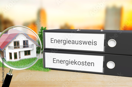 Energieverbrauch. Akten-Ordner beschriftet mit den Wörtern Energieausweis und Energiekosten liegen neben einem Haus-Modell mit Lupe auf einem Schreibtisch. Skyline einer Stadt im Hintergrund.