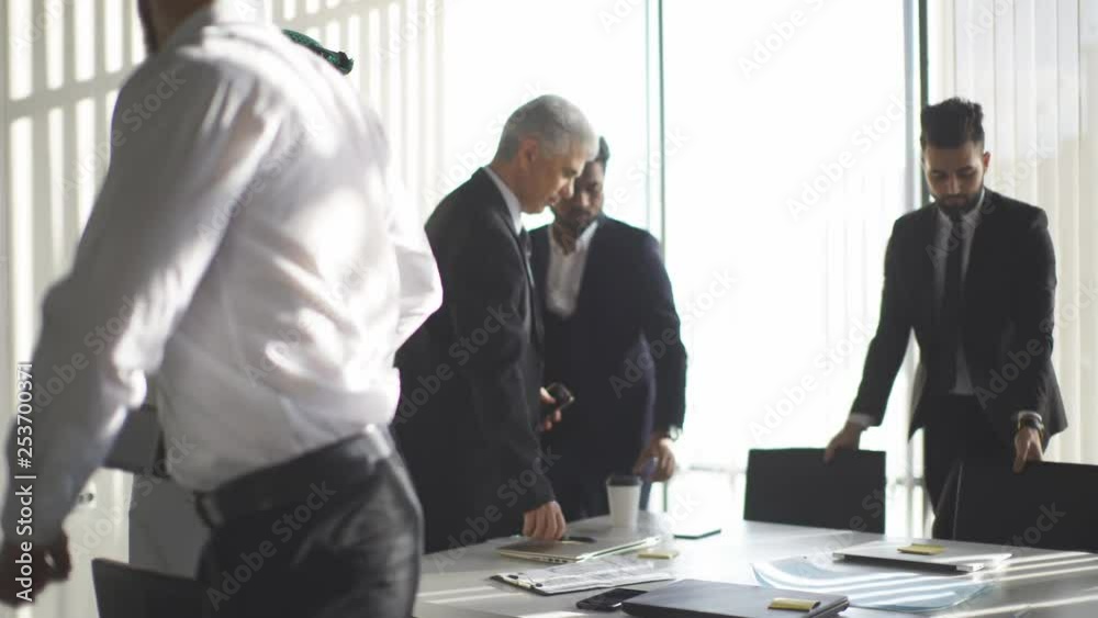 Multi-ethnic male colleagues stand near big windows talking about projects, preparing to Board of directors meeting in modern well-lit conference room with panoramic windows