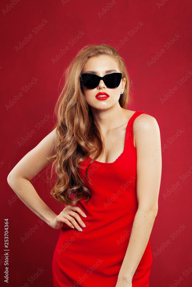 Portrait of young beautiful fashion woman wearing red dress and black sunglasses. Sexy model girl with red lips makeup on red background Portrait of young beautiful fashion woman wearing red dress and black sunglasses. Sexy model girl with red lips makeup on red background