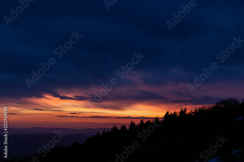 Trees silhouettes against a beautifully colored sky at dusk, with mountains layers in the background © Massimo