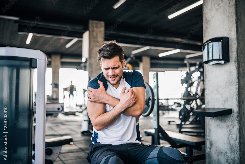 Handsome young sportsman feeling pain in shoulder while working out in the gym.