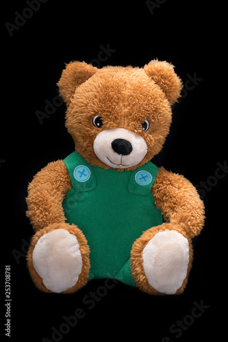 Sitting orange toy Teddy Bear with green overall on black background.