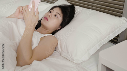 Relaxed asian woman at home reading text message in bright bedroom. charming girl lazy lying in bed stay sleepy on pillow holding cellphone looking screen. young people smart phone addiction.
