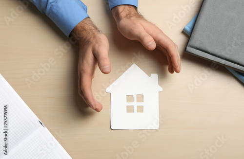 Man holding hands near paper silhouette of house on wooden background, top view. Home insurance