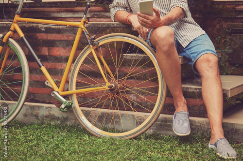 Modern man using cellphone while sitting on the grass with old bicycle.