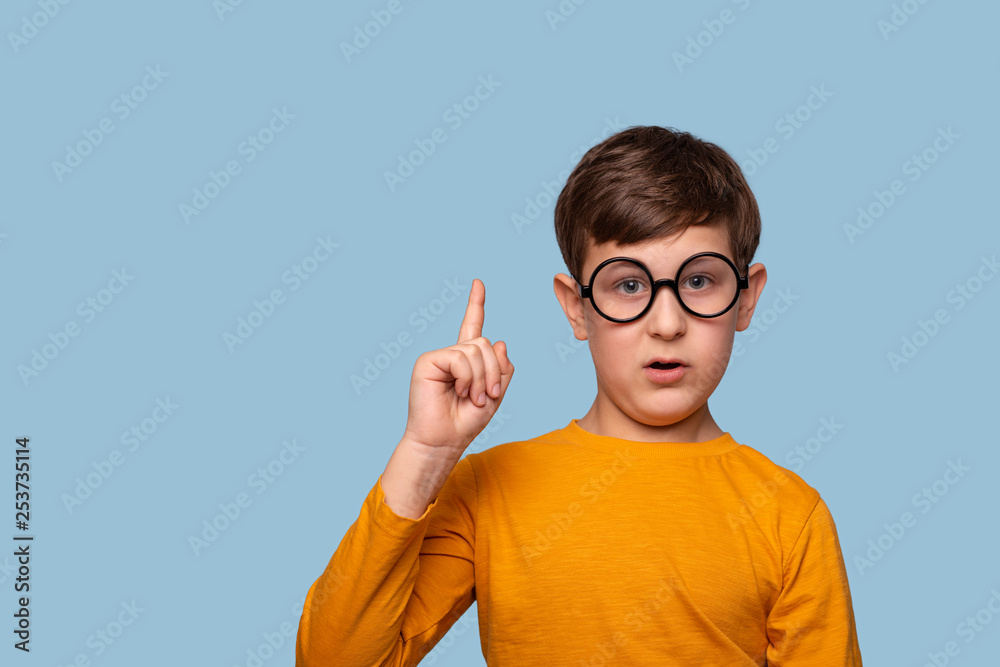 Studio shot of a funny  boy  in round glasses  proving something by raising his index finger up, isolated on blue with copy space