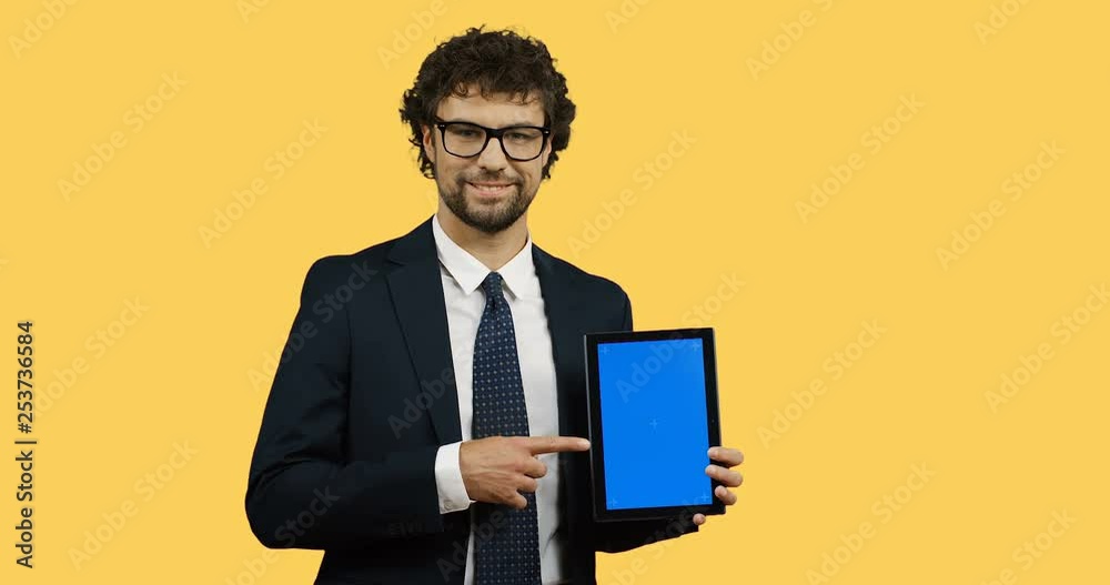 Caucasian good looking man in suit and tie posing with a black tablet computer in hands and pointing with his finger at it on the yellow wall background. Tracking motion Caucasian good looking man in suit and tie posing with a black tablet computer in hands and pointing with his finger at it on the yellow wall background. Tracking motion