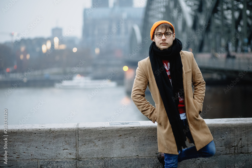 A man stands against the backdrop of a beautiful view. Church of Saint Peter. Cologne Cathedral. Germany. Gothic architecture. Blue jeans, orange hat, beige coat. A man stands against the backdrop of a beautiful view. Church of Saint Peter. Cologne Cathedral. Germany. Gothic architecture. Blue jeans, orange hat, beige coat.