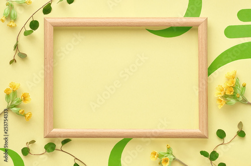 Wooden frame decorated with yellow primrose flowers and leaves, flat lay on yellow paper, copy-space