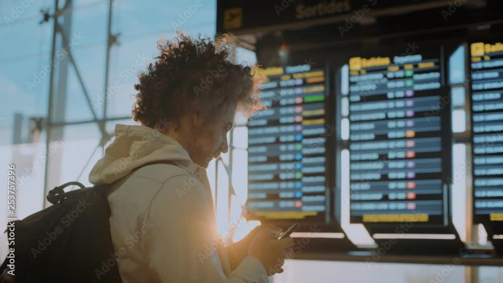Young middle aged traveler man with curly hair and backpack stands in airport departure terminal compares data from email and flight information display system to look for boarding gate or delay.