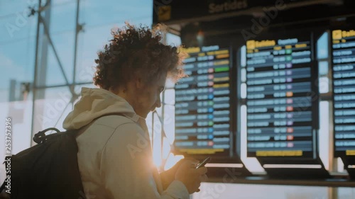 Young middle aged traveler man with curly hair and backpack stands in airport departure terminal compares data from email and flight information display system to look for boarding gate or delay.