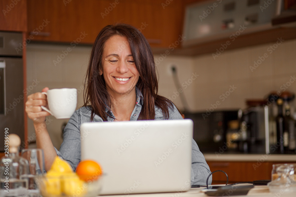Smiling mid adult woman drinking coffee and using laptop in kitchen Smiling mid adult woman drinking coffee and using laptop in kitchen