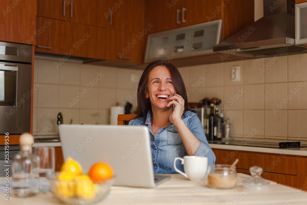 Smiling mid adult woman drinking coffee and using laptop in kitchen Smiling mid adult woman drinking coffee and using laptop in kitchen