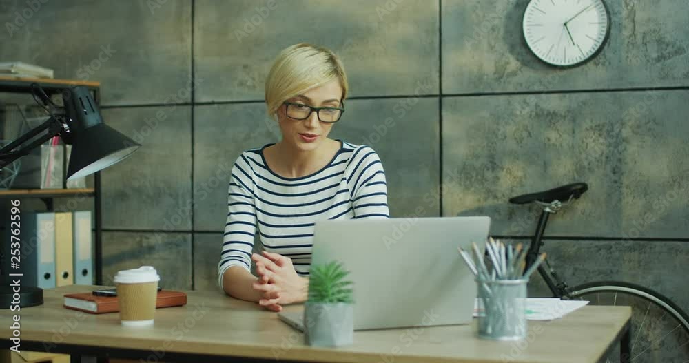 Smiled pretty Caucasian woman sitting at the table in the loft office and talking vis web cam videochat on the laptop computer, demonstrating some charts and explaining something. Smiled pretty Caucasian woman sitting at the table in the loft office and talking vis web cam videochat on the laptop computer, demonstrating some charts and explaining something.
