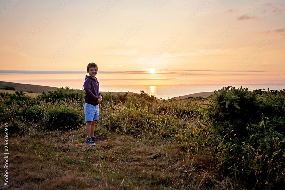 Beautiful preschool boy, standing on the edge of a cliff, contemplating amazing sunset over the ocean