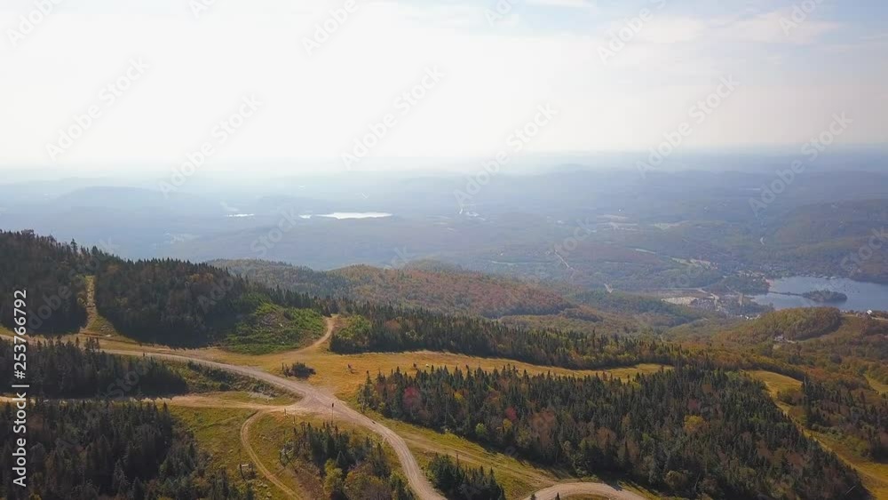 Smooth aerial / drone shot moving forwards over the forest on a hike path during summer season with green, yellow and orange trees, a lake, a small town and mountains on the background.