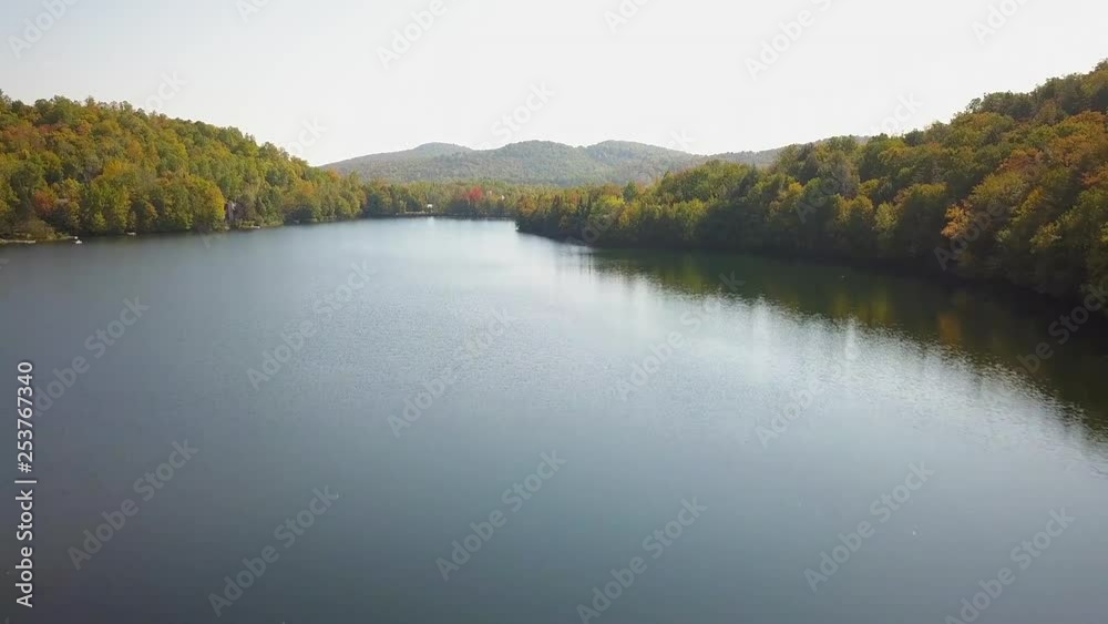 Discovery aerial / drone shot over the lake towards a car racing circuit during summer season with green, yellow and orange trees on the background. Discovery aerial / drone shot over the lake towards a car racing circuit during summer season with green, yellow and orange trees on the background.