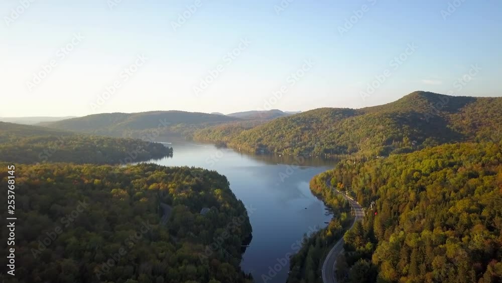 Smooth aerial / drone shot moving forwards over the forest on a driveway, cottages and a lake during summer season with green, yellow and orange trees on the background. Smooth aerial / drone shot moving forwards over the forest on a driveway, cottages and a lake during summer season with green, yellow and orange trees on the background.