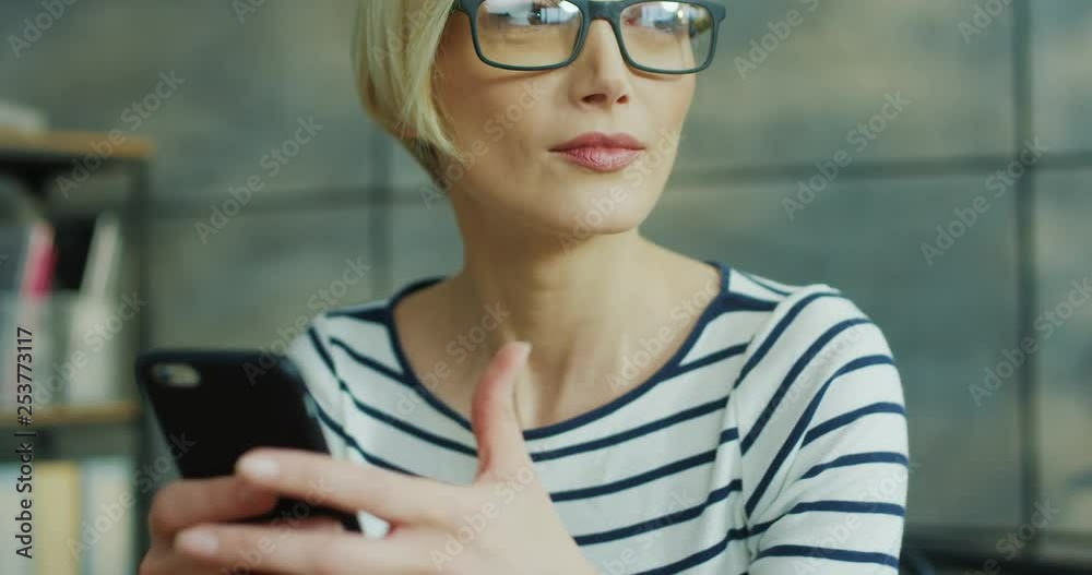 Close up of the pretty blonde woman in glasses tapping and texting on the smartphone with a smile, then looking at the side. Indoors. Close up of the pretty blonde woman in glasses tapping and texting on the smartphone with a smile, then looking at the side. Indoors.