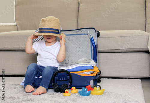 Cute funny little baby boy siiting in blue suitcase with hat on his eyes, packed for vacation full of clothes ready for traveling. Vacation with child. photo