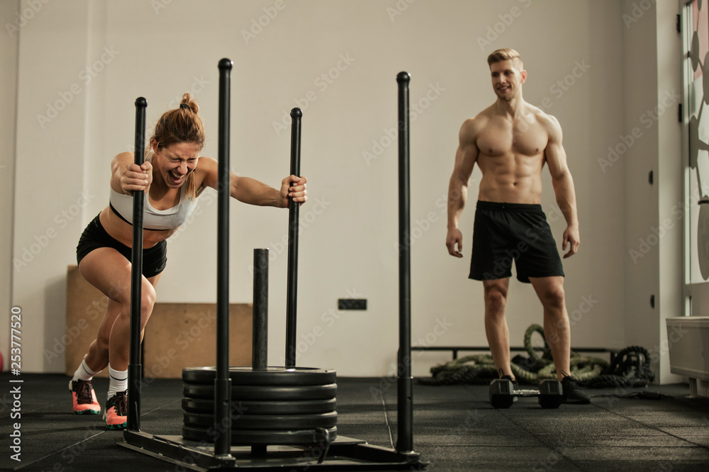 Fototapeta premium Athletic woman making effort while pushing gym sled with weights on cross training.