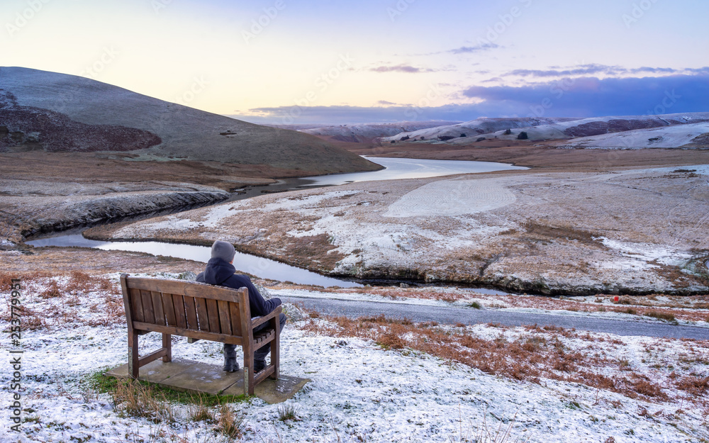Pont Ar Elan, Elan valley, wales. Snowy scene of Afon elan flowing around mountain with hiker sitting on a bench admiring the view