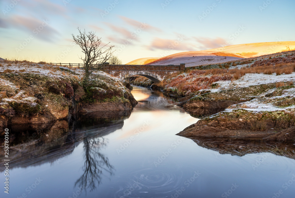 Pont Ar Elan, Elan Valey, wales snowy scene of Afon Elan flowing through a bridge in winter with lone tree reflected in water and early morning sun lighting the top of distant mountains