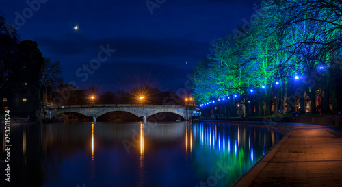 Evesham River Avon night time shot of coloured floodlight on trees being reflected in a still water with Workman Bridge, moon and stars.